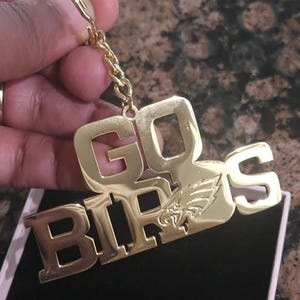 Può includere: Un portachiavi color oro con le parole "GO BIRDS" in grassetto maiuscolo. La parola "BIRDS" presenta il logo dei Philadelphia Eagles. Il portachiavi è attaccato a una catena e un anello dorati.