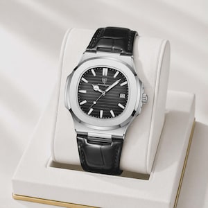 May include: A silver-toned wristwatch with a black dial and a black leather band. The watch features a date window and a textured dial. The watch is displayed on a white stand.