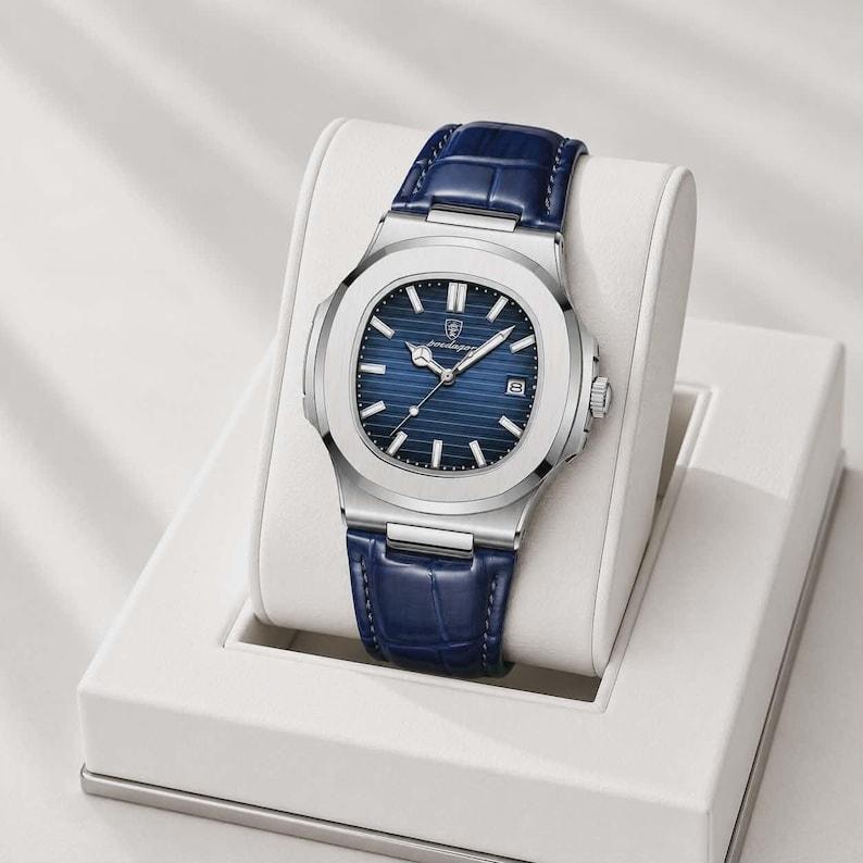 May include: A silver-toned wristwatch with a blue dial and a blue leather strap. The watch has a rectangular face with a date display and is presented on a white display stand. The watch band is approximately 8 inches long.