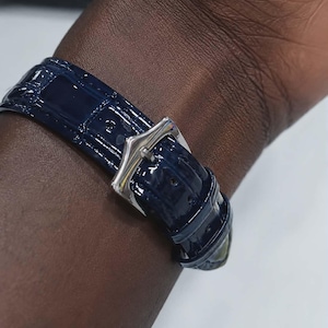 May include: A close-up of a navy blue watch band with a silver buckle. The band has a glossy, textured finish and is secured on a wrist. The watch band is the primary focus of the image.