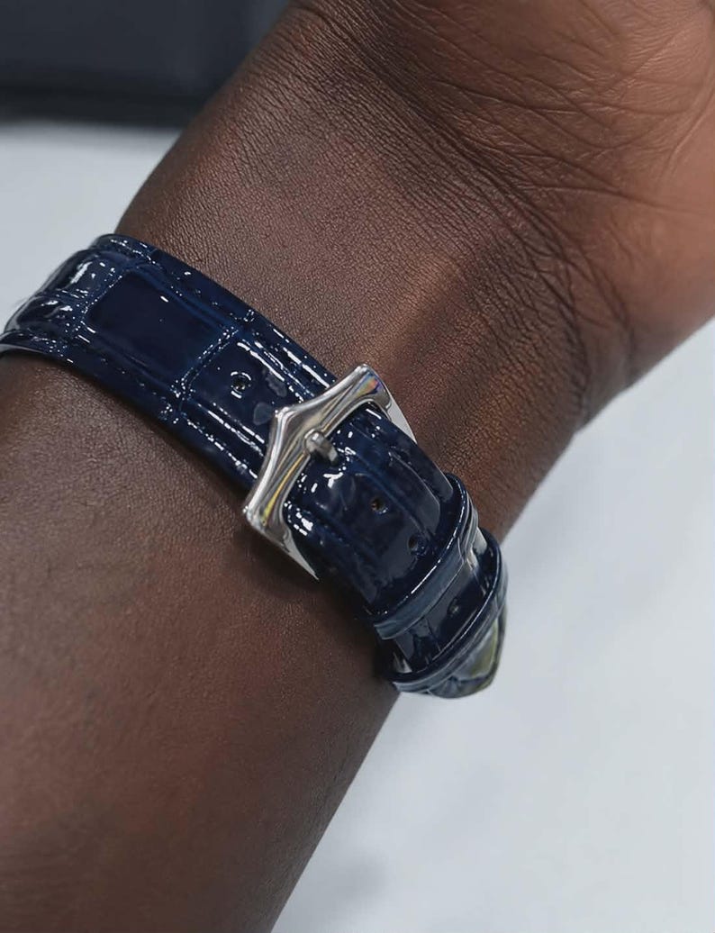May include: A close-up of a navy blue watch band with a silver buckle. The band has a glossy, textured finish and is secured on a wrist. The watch band is the primary focus of the image.