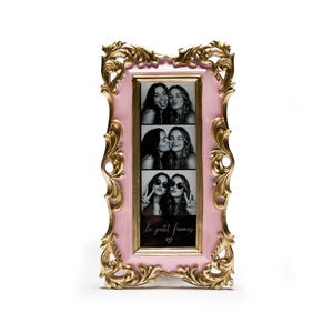 May include: A pink and gold ornate picture frame with three black and white photo strips. The frame has a rectangular opening and features the text "le petit frames" with a heart symbol. The frame is on a white background.