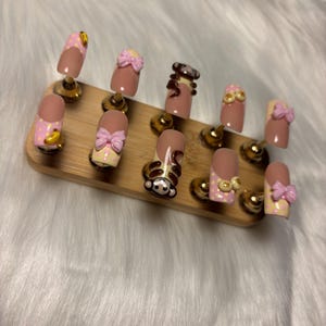 May include: A collection of artificial nails on a wooden stand. The nails showcase diverse designs, such as pink and yellow polka dots, bows, and a monkey. Some nails have a nude base with detailed nail art.