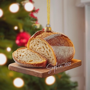 May include: A bread-shaped Christmas ornament with a slice cut out, resting on a small wooden cutting board. The ornament is suspended by a gold string. The name "Sophia 2025" is written on the board in cursive.