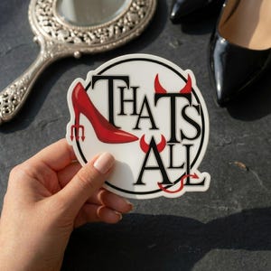 May include: A white sticker with the text "THAT'S ALL" in black, with red devil horns and a red high-heeled shoe. The sticker is held by a hand, with a mirror and black high heels in the background.