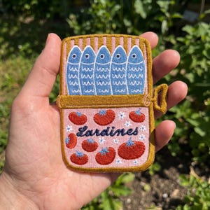May include: An embroidered patch shaped like a rectangular tin can with a gold border. The top section shows five blue fish, while the bottom pink section features red tomatoes, small white flowers, and the dark blue text Lardines.