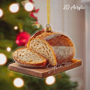 May include: A 2D acrylic ornament shaped like a loaf of sourdough bread on a wooden cutting board. The bread has a golden-brown crust and an airy interior. The text "Sophia 2025" is printed on the board. It hangs from a gold string.
