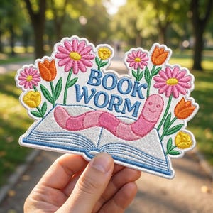 May include: Embroidered patch featuring a smiling pink worm on an open book, surrounded by pink daisies, yellow flowers, and orange tulips. The words "BOOK WORM" are in blue. The patch is held in a hand against a blurred green background.