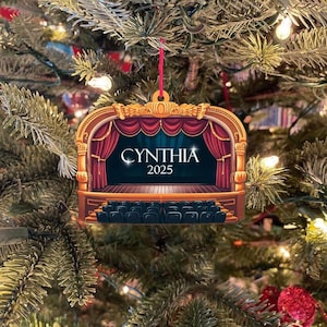May include: A theater-themed Christmas ornament with a red string for hanging. The ornament features a stage design with red curtains, a stage, and seating. The name "CYNTHIA" and the year "2025" are displayed on the stage.