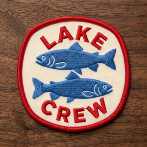 May include: A circular embroidered patch with a cream-colored background and a red stitched border. The patch features two blue fish stacked horizontally in the center, with the red embroidered text LAKE above and CREW below the fish.