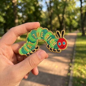 May include: Embroidered patch featuring a green and yellow caterpillar with the text "EAT THE RICH". The caterpillar has a red head with blue eyes and black antennae. The patch is held in a hand against a blurred outdoor background.