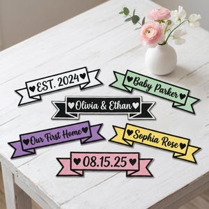May include: Six rectangular fabric banners with pointed ends, each featuring black text and heart icons. The banners are white, light green, black, purple, yellow, and pink, displaying custom phrases like EST. 2024, Baby Parker, Olivia & Ethan, Our First Home, Sophia Rose, and 08.15.25.