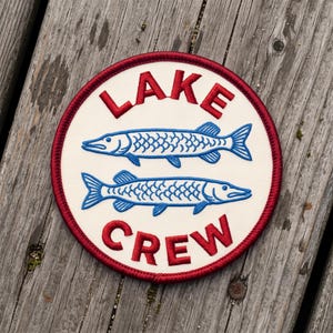 May include: A circular embroidered patch with a cream-colored background and a dark red stitched border. The text "LAKE" is embroidered in red at the top, and "CREW" is in red at the bottom. Two blue fish are centered between the text.