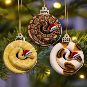 May include: Three Christmas ornaments featuring coiled snakes in shades of brown, yellow, and white, each wearing a Santa hat. The ornaments are round with a gold string for hanging and the year "2025" printed on top. Each snake ornament has the name "Bella" on the hat.