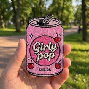 May include: A pink patch in the shape of a soda can with the words "Girly Pop" in white script. The patch features two cherries, stars, and the text "12 FL OZ."