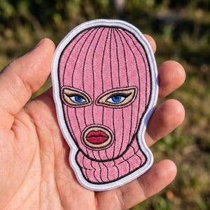 May include: An embroidered patch featuring a pink balaclava mask with vertical ribbed texture. The mask has cutouts revealing bright blue eyes with black eyeliner and dark pink lips. The entire design is outlined in a thick white border.