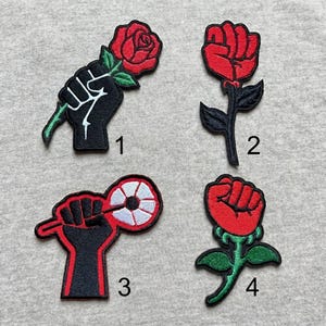 May include: Four embroidered patches with red and black designs. The designs include a fist holding a red rose, a red fist with a black stem and leaves, a fist holding a red and white design, and a red fist with a green stem and leaves.