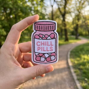 May include: A pink and white embroidered patch in the shape of a pill bottle. The patch features the words "CHILL PILLS" in white lettering on a white background. The bottle is filled with pink and white pills.