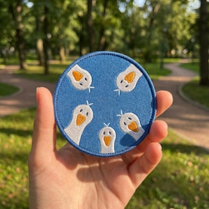 May include: A round, blue felt coaster featuring a design of five white geese with orange beaks. The geese are arranged around the center of the coaster. The coaster is held in a hand against a blurred green background.