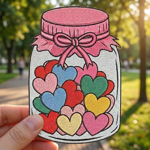 May include: Embroidered patch featuring a jar filled with colorful hearts. The jar has a pink lid and a pink bow. The hearts are various colors, including red, blue, green, yellow, and pink. The patch is held in front of a blurred outdoor background.