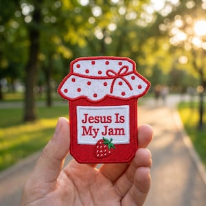 May include: A red and white embroidered patch in the shape of a jam jar. The patch features the text "Jesus Is My Jam" and a small strawberry illustration. The jar has a white lid with red polka dots and a red bow.