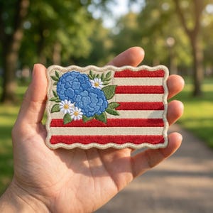 May include: Embroidered patch featuring a stylized American flag design. The flag has red and white stripes with a blue floral arrangement in the upper left corner. The flowers are blue hydrangeas and white daisies with green leaves.