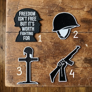 May include: Four black and white patches on a wooden surface. The patches include a silhouette with the text "FREEDOM ISN'T FREE BUT IT'S WORTH FIGHTING FOR", a military helmet, a cross with a helmet, and a hand holding a rifle.