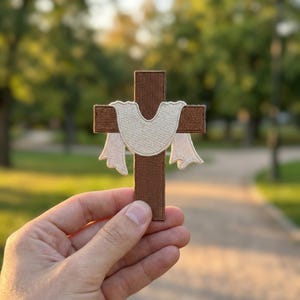 May include: A brown cross patch with a cream-colored cloth draped over it. The patch has a detailed embroidered design. The background is blurred, with green trees and a pathway visible.