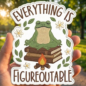 May include: Embroidered patch with the text "EVERYTHING IS FIGUREOUTABLE". The patch features a green frog meditating on a stack of books, with a campfire at the bottom. White flowers and green leaves surround the text and frog.