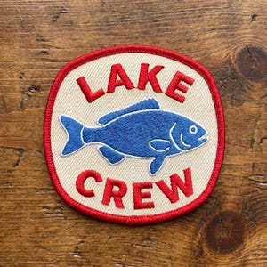 May include: Embroidered patch with a cream-colored background, red border, and red text that reads "LAKE CREW." A blue fish is in the center. The patch is circular with a slightly rectangular shape.