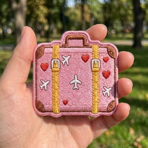 May include: Pink embroidered patch in the shape of a suitcase. The patch features yellow straps, brown corners, and is decorated with red hearts and white airplanes. A small airplane and a larger airplane are embroidered on the front.