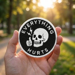 May include: Round black and white embroidered patch with the words "EVERYTHING HURTS" surrounding a skull and a thumbs-up gesture with a lightning bolt. The patch is held in a hand against a blurred green background.
