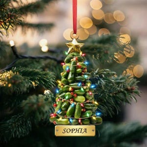 May include: A unique Christmas tree ornament crafted from green pickles, adorned with colorful lights and a gold star topper. The ornament hangs from a red ribbon and features the name "SOPHIA" on a gold plaque. Festive and whimsical.