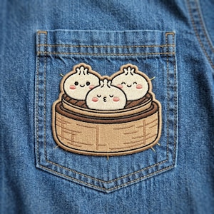 May include: An embroidered patch featuring three cream-colored bao buns with simple black facial features and pink cheeks, resting inside a light brown bamboo steamer basket, stitched onto the pocket of a blue denim shirt.