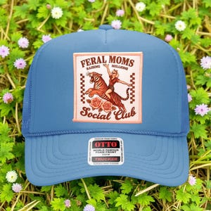 Puede incluir: Gorra de camionero azul claro con parte trasera de malla y frontal de espuma. El parche frontal dice "FERAL MOMS RAISING HELLIONS SOCIAL CLUB" con un gráfico de una mujer montada en un tigre. La gorra tiene un cierre a presión.