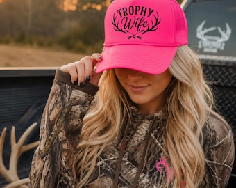 Könnte beinhalten: Eine leuchtend neonpinke Trucker-Cap mit einem schwarzen Grafikdruck auf der Vorderseite. Das Design zeigt den Schriftzug Trophy Wife in fetter Schrift über zwei gekreuzten Pfeilen und stilisiertem Hirschgeweih mit einem kleinen Herz.