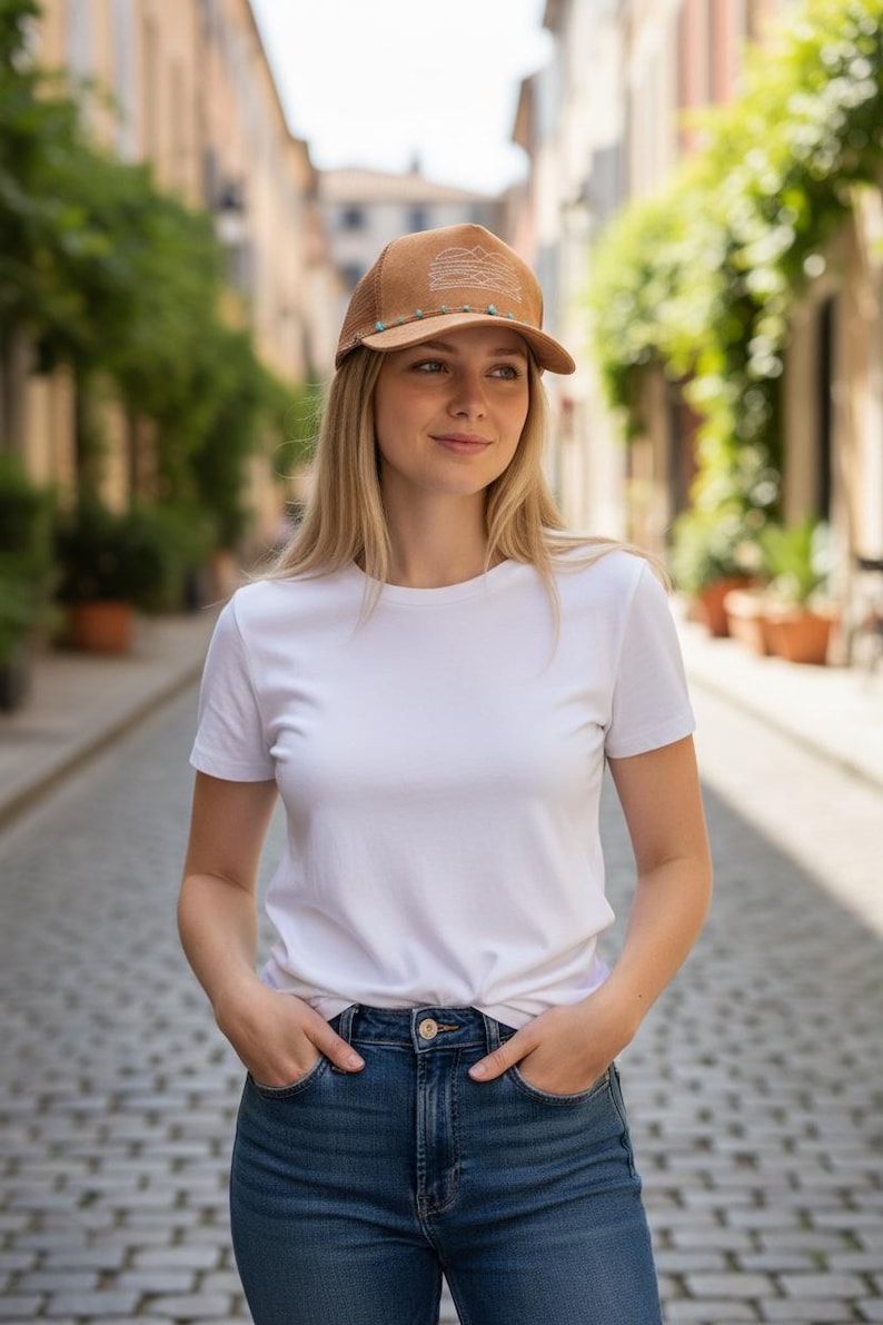 K&ouml;nnte beinhalten: Braune Trucker-M&uuml;tze mit Bergmotiv und t&uuml;rkisfarbenen Perlen. Die M&uuml;tze wird mit einem wei&szlig;en T-Shirt und einer blauen Jeans getragen. Das Bild wird im Freien auf einer Kopfsteinpflasterstra&szlig;e aufgenommen.