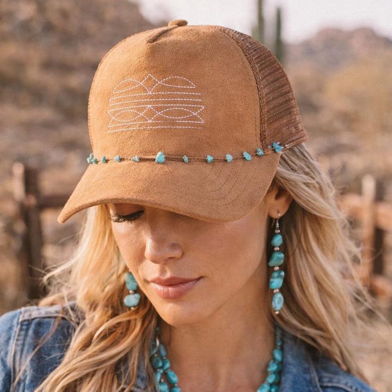 K&ouml;nnte beinhalten: Braune Trucker-Cap mit Wildlederfront und Mesh-R&uuml;ckseite. Sie hat ein wei&szlig;es Stickmuster und kleine t&uuml;rkisfarbene Steine am Rand. Die Kappe ist Teil einer Kollektion im Western-Stil mit rustikaler &Auml;sthetik.