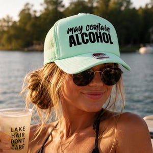 Könnte beinhalten: Hellgrüne Trucker-Cap mit dem Text "may contain ALCOHOL" in Schwarz. Die Kappe wird von einer Person mit Sonnenbrille getragen, die einen durchsichtigen Plastikbecher mit dem Text "LAKE HAIR DON'T CARE" hält. Im Hintergrund sind See und Bäume zu sehen.