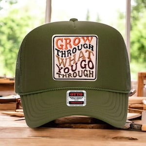 May include: Olive green trucker hat with a mesh back and a curved brim. The front patch reads "GROW THROUGH WHAT YOU GO THROUGH" in orange and brown lettering. The hat also has a small Otto tag.