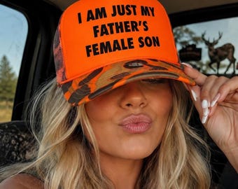 Könnte beinhalten: Eine leuchtend neonorange Trucker-Cap mit Camouflage-Schirm und Netzrückseite. Auf der Vorderseite steht in schwarzer, fetter serifenloser Schrift der Text "I AM JUST MY FATHER'S FEMALE SON". Die Kappe hat eine strukturierte Krone und einen gebogenen Schirm.
