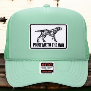 Könnte beinhalten: Hellgrüne Trucker-Cap mit Mesh-Rückseite und gebogenem Schirm. Auf der Vorderseite befindet sich ein rechteckiger Aufnäher mit einer schwarz-weißen Hundeillustration und dem Text "POINT ME TO THE BAR". Ein kleines weißes Etikett ist vorhanden.