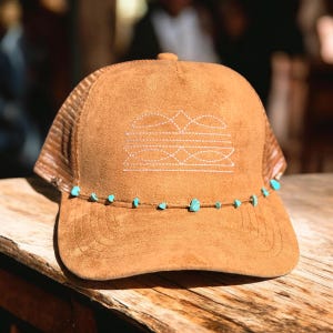 K&ouml;nnte beinhalten: Braune Trucker-Cap mit Wildleder-&auml;hnlicher Textur. Die Vorderseite hat wei&szlig;e, gestickte geometrische Designs. Der Rand ist mit kleinen t&uuml;rkisfarbenen Perlen verziert. Die Netzr&uuml;ckseite ist hellbraun.