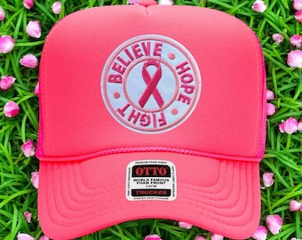 Könnte beinhalten: Eine leuchtend pinkfarbene Trucker-Mütze mit Mesh-Rückseite. Auf der Vorderseite befindet sich ein kreisförmiges Design mit einem rosa Band und den Worten "Believe, Hope, Fight". Die Mütze hat einen Schnappverschluss und ein Etikett "World Famous Foam Front".