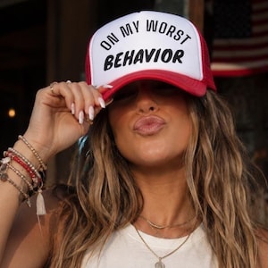 May include: A white and red trucker hat with the text "ON MY WORST BEHAVIOR" in black. The hat is worn by a person with long, wavy hair and multiple bracelets. An American flag is visible in the background.