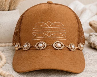 Könnte beinhalten: Eine beigefarbene Baseballkappe aus Wildleder mit Netzrücken und gebogenem Schirm. Das Vorderteil zeigt weiße, gestickte geometrische Linien. Eine goldfarbene Kette, verziert mit kleinen, runden, weißen Kunstperlen, umgibt die Basis der Kappe.