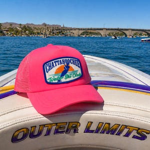 May include: A bright pink trucker hat with a mesh back and a patch that reads "Chattahoochee Water Ski Club." The hat is resting on the white seat of a boat with the words "Outer Limits" in gold lettering. The background shows a lake, bridge, and blue sky.
