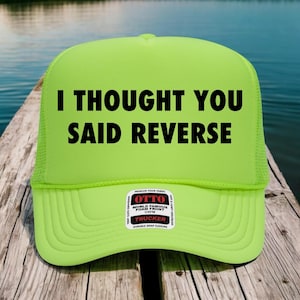 May include: A bright green trucker hat with the words "I THOUGHT YOU SAID REVERSE" printed in black. The hat features a foam front, mesh back, and a snap closure. A small white label with black text is visible on the front.