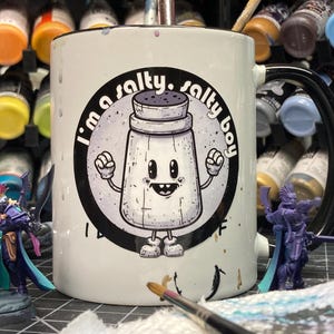 May include: White ceramic mug with a black rim and handle, featuring a cartoon salt shaker illustration with the text "I'm a salty. salty boy" in a circular design. Miniature figurines and paint bottles are in the background.