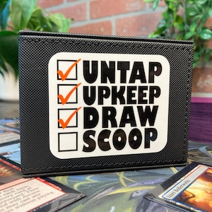 May include: Black card storage box with a white sticker that reads "UNTAP UPKEEP DRAW SCOOP" with orange check marks. The box is surrounded by playing cards and a stack of purple cards.